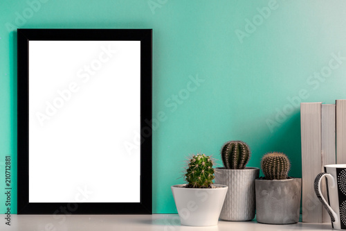 Fototapeta Naklejka Na Ścianę i Meble -  Blank black frame with cactuses, books and a black mug with coffee on the background of a bright mint wall