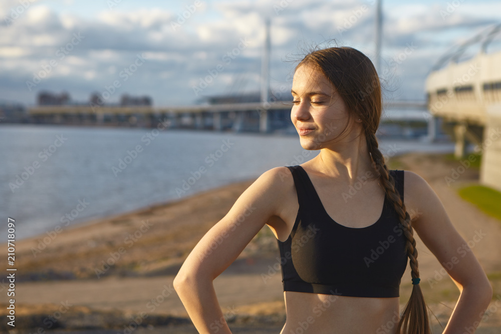 Waist up shot of attractive gorgeous young sportswoman with athletic slim body enjoying warm summer sun during morning training outdoors, smiling and closing eyes, vast river in background