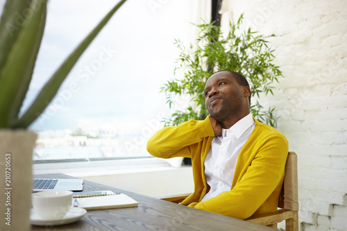 Indoor shot of stylish Afro American male interior designer wearing yellow cardigan over white shirt sitting at wooden desk in modern office with brick wall and green plants, having pensive look photo