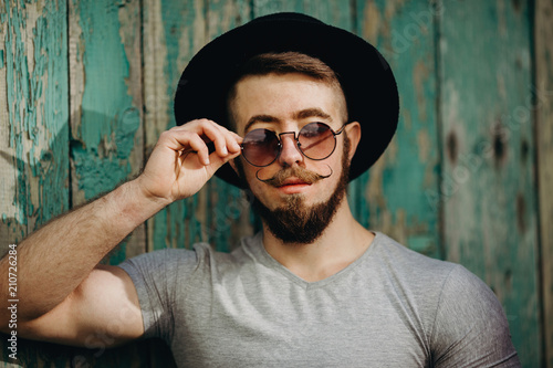 Close up of stylish hipster man in hat and sunglasses wearing fashionable mustaches. Fashion, people, lifestyle concept