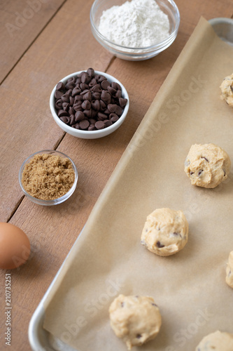 Row of chocolate chip cookie ingredients in bowls, beside dough balls on parchment paper