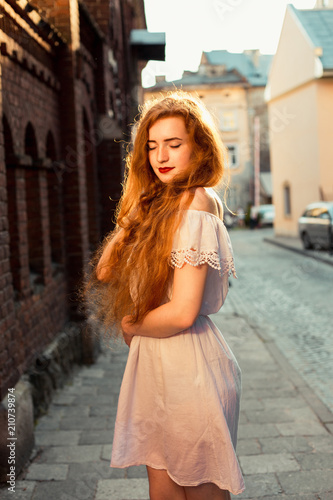 Romantic red haired model with long curly hair wearing white dress posing in evening sunlight