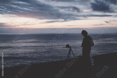 young hipster guy with long hair takes a picture on photo of a sea sunset at night on a sunset background, silhouette photographer using camera in travel,