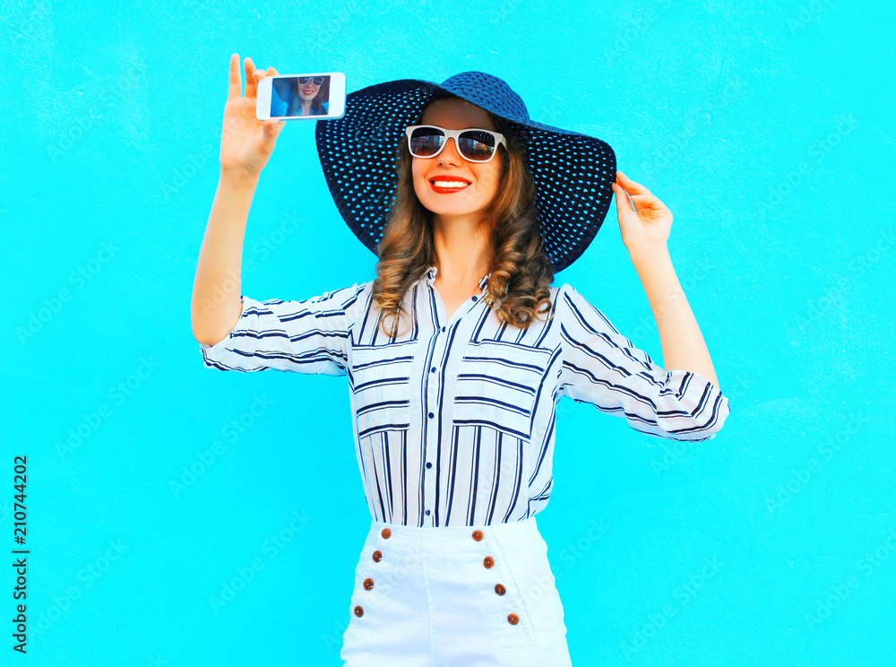 Cool girl is taking a picture on a smartphone wearing a straw summer hat, white pants over colorful blue background Cool girl is taking a picture on a smartphone wearing a straw summer hat, white pants over colorful blue background