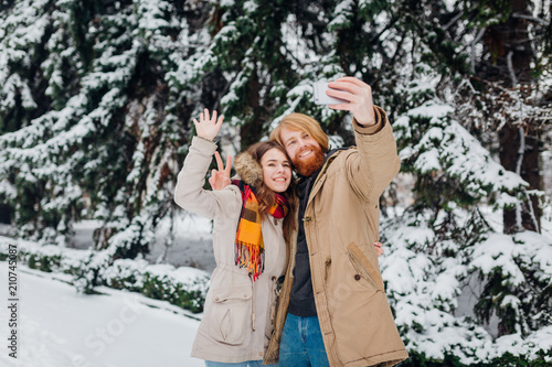Young couple in love make a joint photo of themselves on a mobile phone. The guy holds a smartphone on his elongated hand and makes a photo, waving hands, a greeting gesture against park in winter