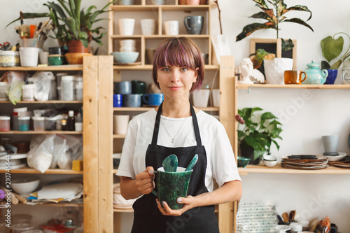 Pretty girl with colorful hair in black apron and white T-shirt holding handmade bowl in hands dreamily looking in camera spending time at modern pottery studio photo