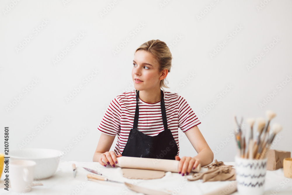 Beautiful girl in black apron and striped T-shirt sitting at the table holding rolling pin working with clay and dreamily looking aside at pottery studio