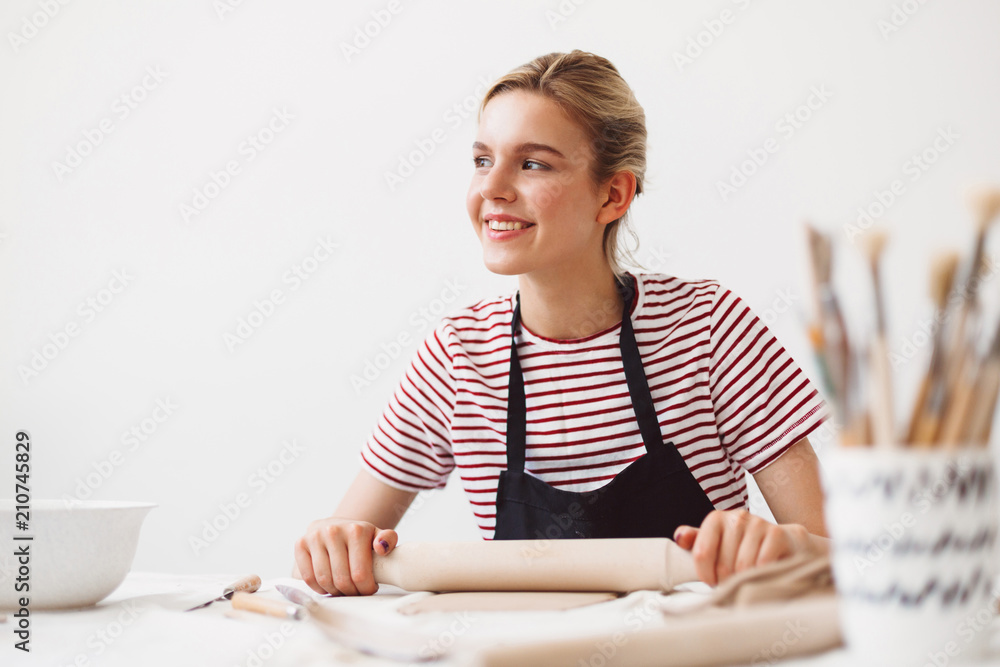 Smiling girl in black apron and striped T-shirt sitting at the table with rolling pin and working with clay happily looking aside at pottery studio Smiling girl in black apron and striped T-shirt sitting at the table with rolling pin and working with clay happily looking aside at pottery studio