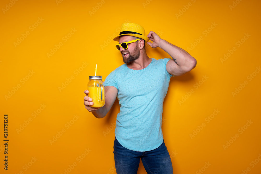 Creative male in sunglasses and summer clothes in yellow and blue colors standing relaxed with hand touching stylish hat and drinking from creative cocktail bottle on yellow studio background.