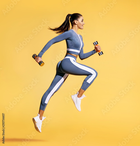 Sporty woman jumping with dumbbells. Photo of active woman in sportswear on yellow background. Dynamic movement. Side view. Sport and healthy lifestyle