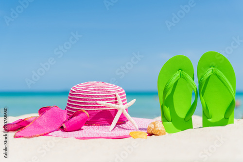 Beach accessories including flip flop, starfish, beach hat and sea shell on sandy beach, green sea and blue sky background for summer holiday and vacation concept.