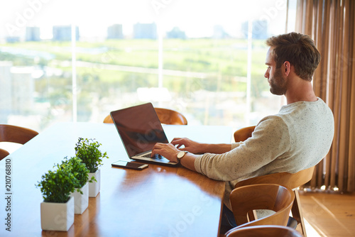 modern day white man busy working on his notebook while seated at his dining table at home, with a gorgeous view of the city through the large glass windows.