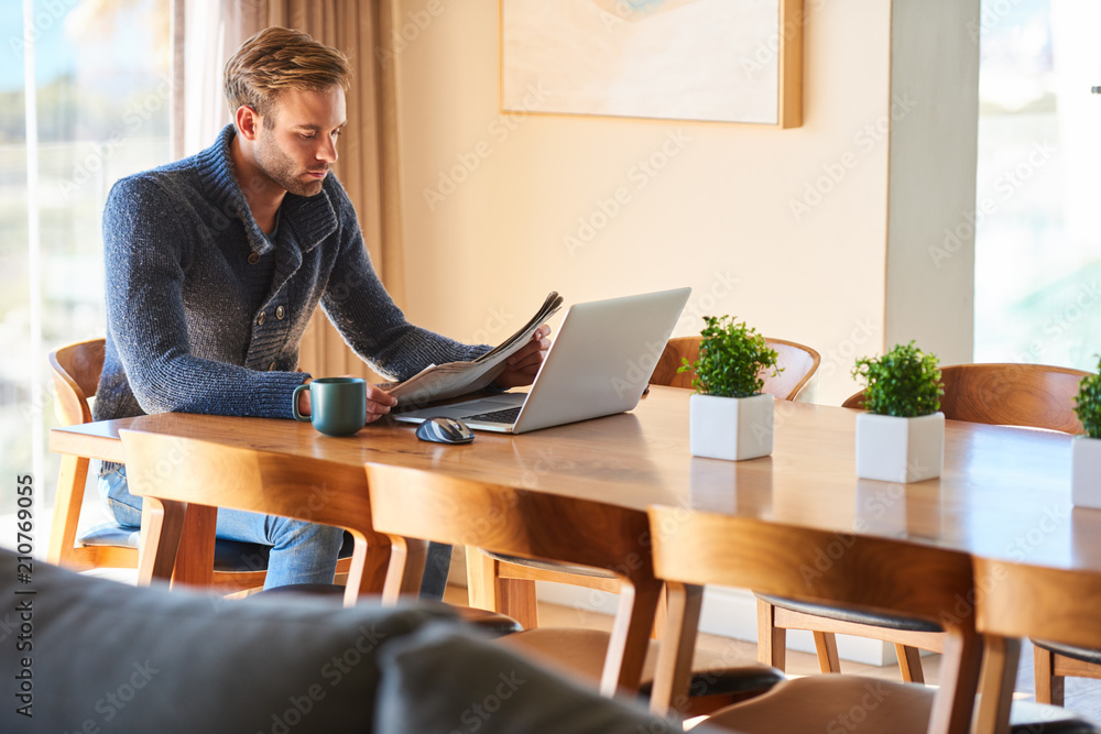 Sophisticated modern man sitting at his dining table with his morning cup of coffee at his side and his notebook ready for work in front of him while reading the daily newspaper. Sophisticated modern man sitting at his dining table with his morning cup of coffee at his side and his notebook ready for work in front of him while reading the daily newspaper.