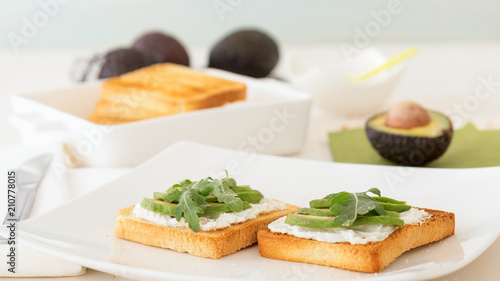 Roasted toasts with cream cheese and slices of avocado, leafs of rucola and pink Himalayan salt. Front view, low angle, focus on the centre of both breads. White wood background.