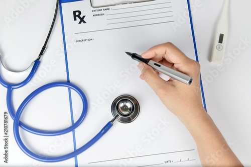 Top view of a doctor's hand writing a prescription. A stethoscope and thermometer are also on white background.