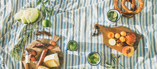 Flat-lay of summer picnic set with fruit, cheese, sausage, bagels and lemonade over striped blanket, top view, copy space