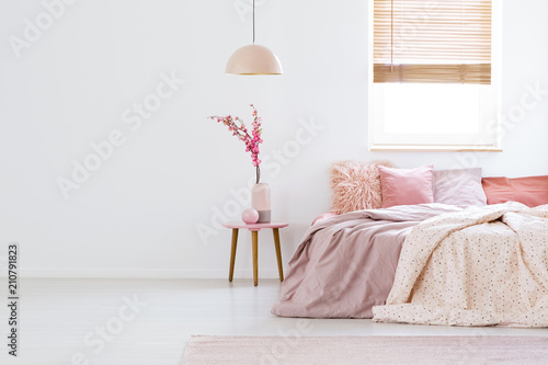 Lamp above table with flowers in pink pastel bedroom interior with window above bed. Real photo photo