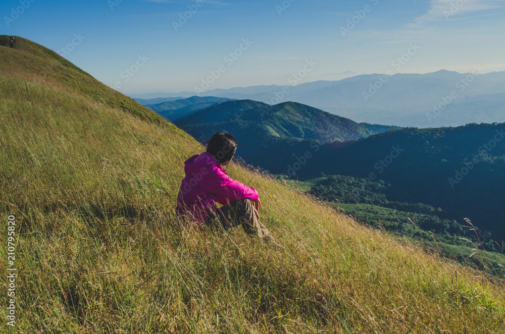Young traveling woman wearing hat and sitting on the top of the mountain cliff with relaxing mood and watching beautiful view of woods and blue sky and clouds on vacation