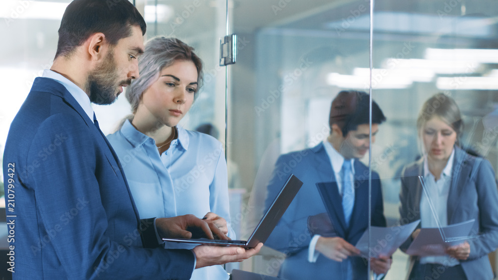 Businessman Holding Laptop Talks With Businesswoman in the Glass of the Office, Discussing Business Related Issues. Busy Business People Walking Through the Hallway of the Corporate Building.