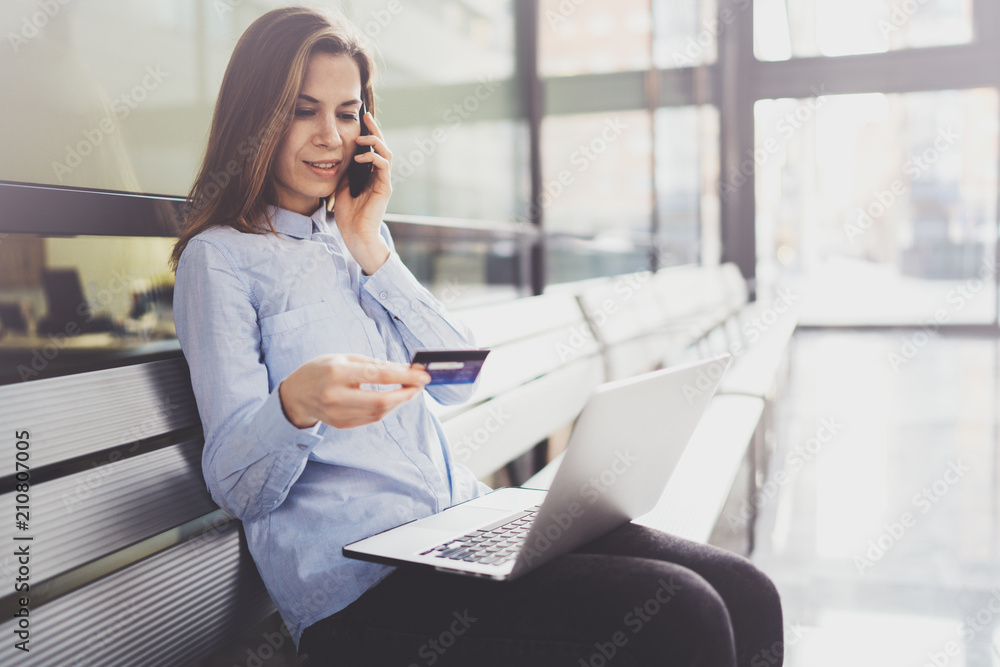 Young attractive businesswoman holding credit card in female hand and using smartphone and mobile laptop while sitting at business center.Blurred background. Young attractive businesswoman holding credit card in female hand and using smartphone and mobile laptop while sitting at business center.Blurred background.