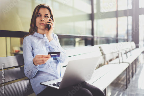 Young attractive businesswoman holding credit card in female hand and using smartphone and mobile laptop while sitting at business center.Blurred background.