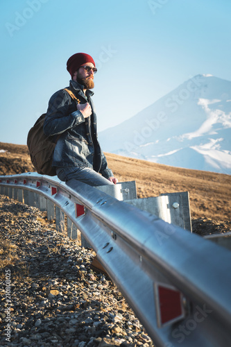 Bearded tourist hipster man in sunglasses with a backpack sitting on a roadside bump and watching the sunset against the background of a snow-capped mountain