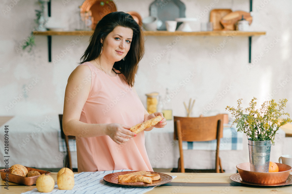 Young beautiful housewife in dress having breakfast on kitchen. Lovely woman posing and dreaming in country french style decorated interior zone. Rustic dining room with fresh food and tableware.