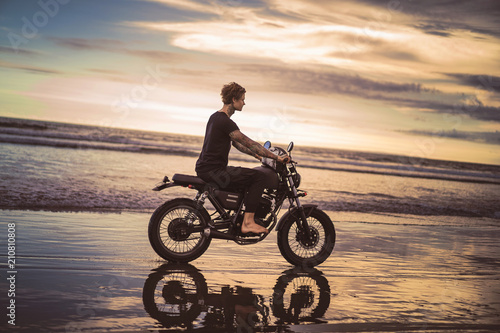 side view of tattooed man riding motorbike on ocean beach during beautiful sunrise