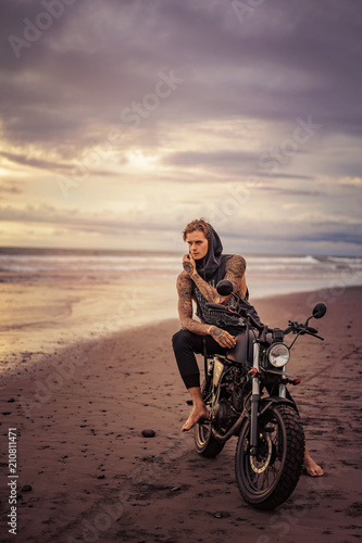 handsome tattooed biker posing on motorcycle on ocean beach