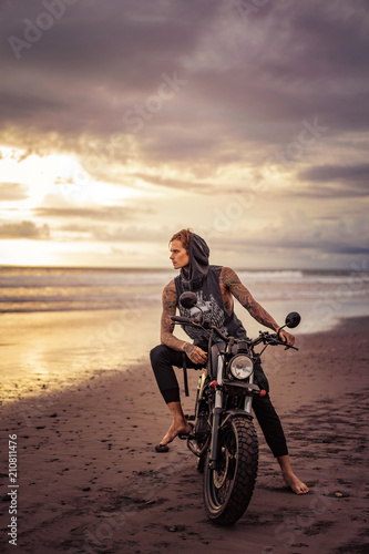handsome tattooed man sitting on motorcycle on ocean beach and looking away