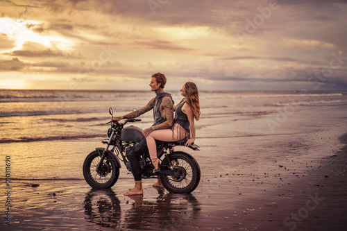tattooed boyfriend and girlfriend sitting on motorcycle at beach during sunrise