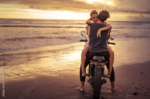 heterosexual couple hugging on motorcycle on ocean beach during beautiful sunrise