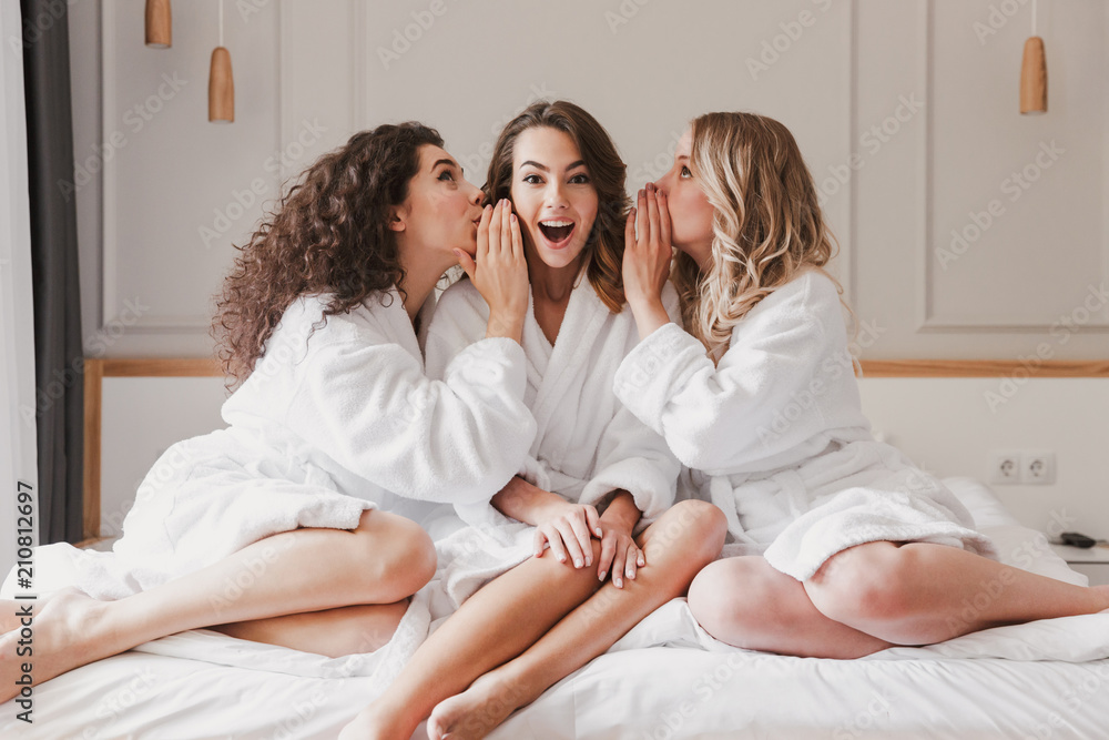 Image of young three women 20s wearing bathrobe sitting on big bed, and telling gossips to each others during bridal shower in cozy apartment or fashionable hotel room - obrazy, fototapety, plakaty 