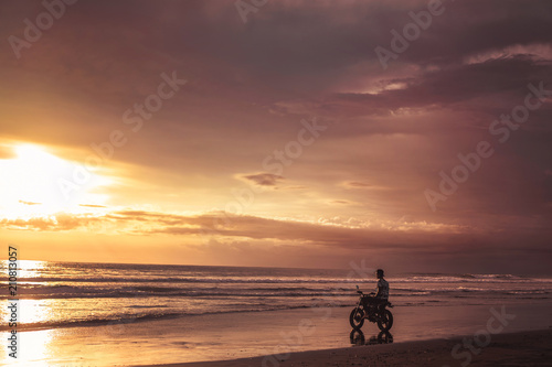man sitting on motorbike on seashore and looking at beautiful sunset