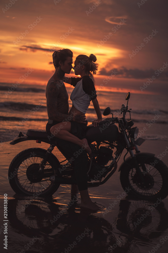 Fototapeta passionate couple hugging and going to kiss on motorcycle at beach during sunset