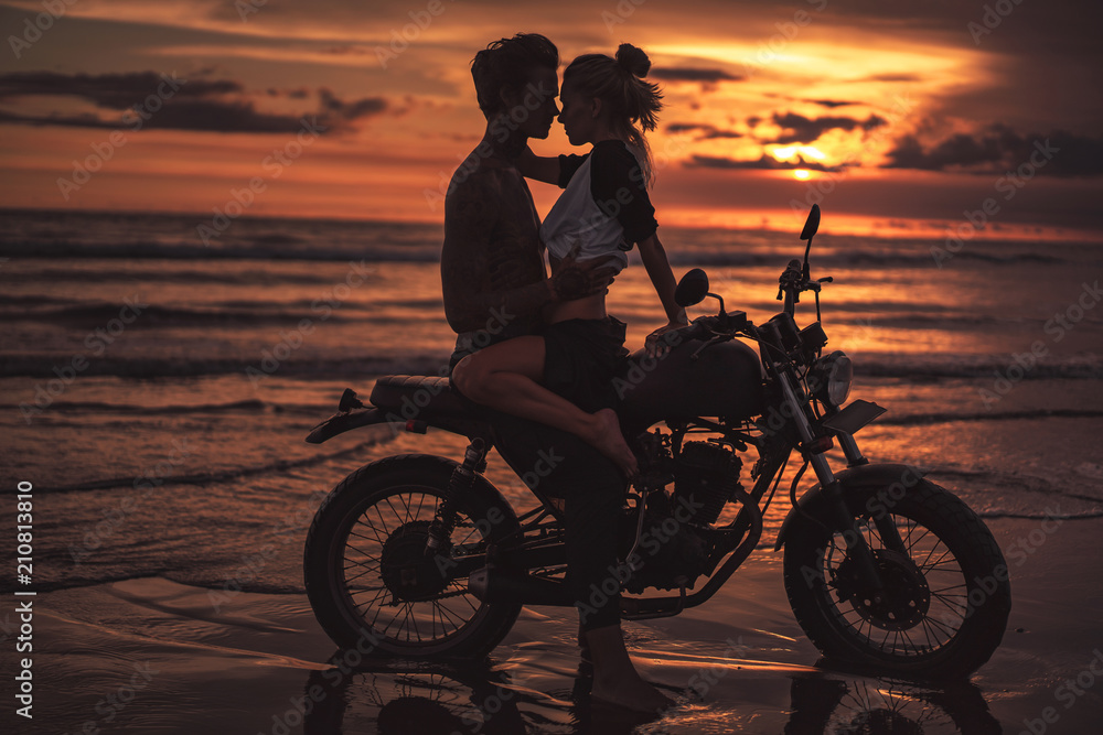 Fototapeta affectionate couple hugging and going to kiss on motorcycle at beach during sunset