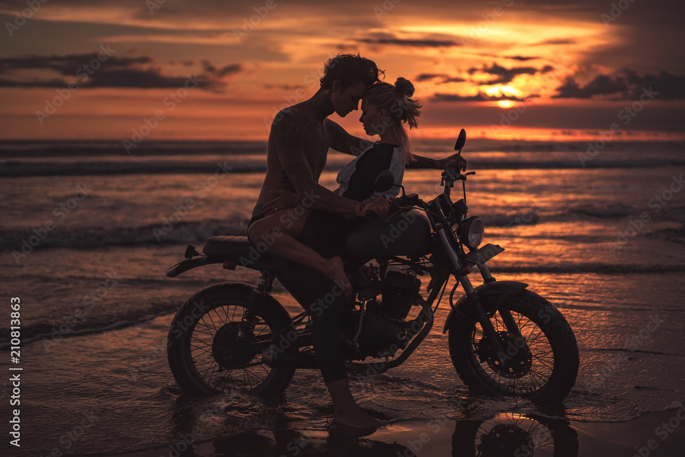 Fototapeta passionate couple hugging and touching with foreheads on motorcycle at beach during sunset