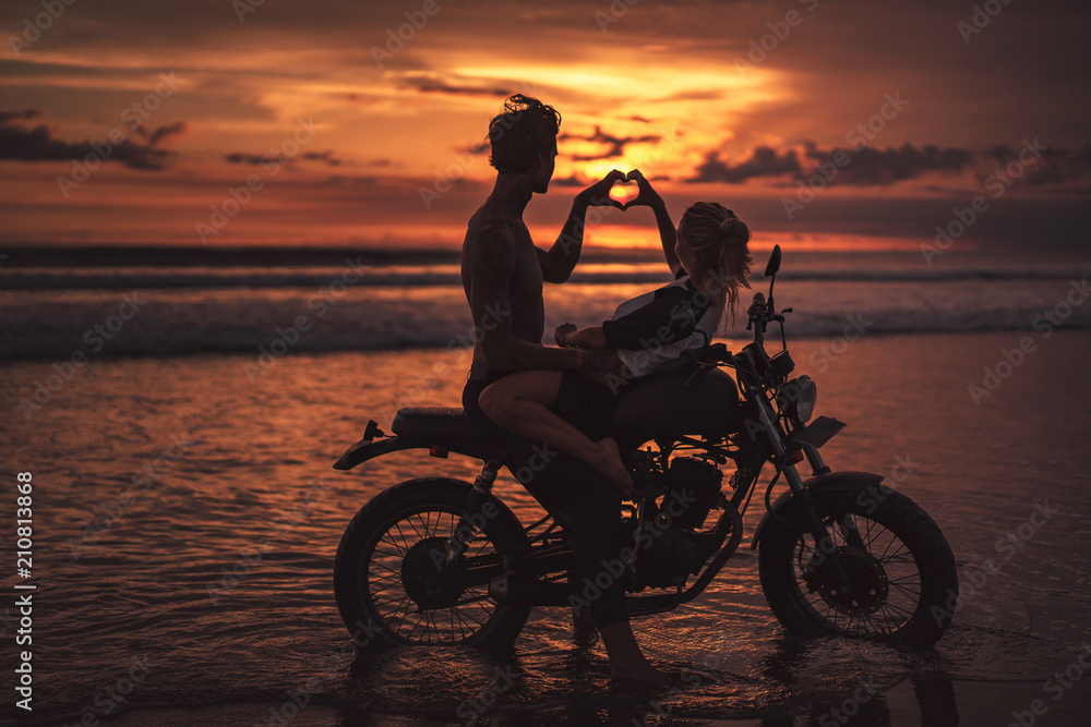 Fototapeta premium side view of couple making heart with fingers on motorcycle at beach during sunset