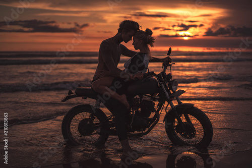 passionate couple hugging and touching with foreheads on motorcycle at beach during sunset