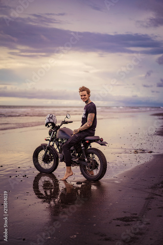 smiling handsome tattooed man sitting on motorbike on ocean beach during sunrise