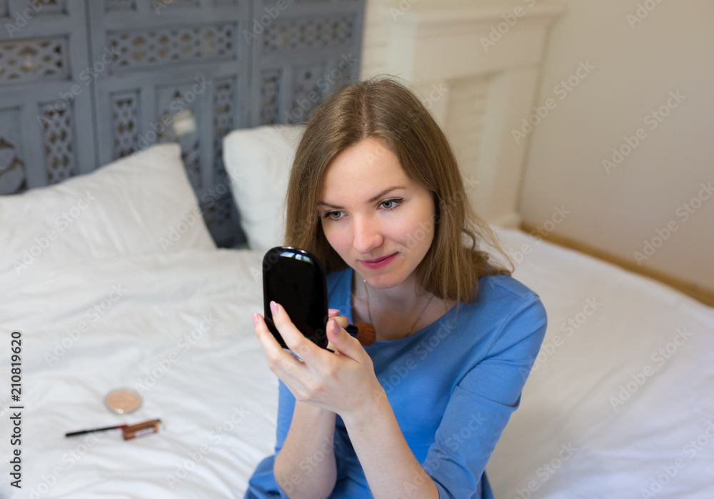 Beautiful girl in blue dress is sitting on white bed linen in minimalist loft design room. Young woman is doing make up. She is looking in the mirror and powders her face. Beautiful girl in blue dress is sitting on white bed linen in minimalist loft design room. Young woman is doing make up. She is looking in the mirror and powders her face.