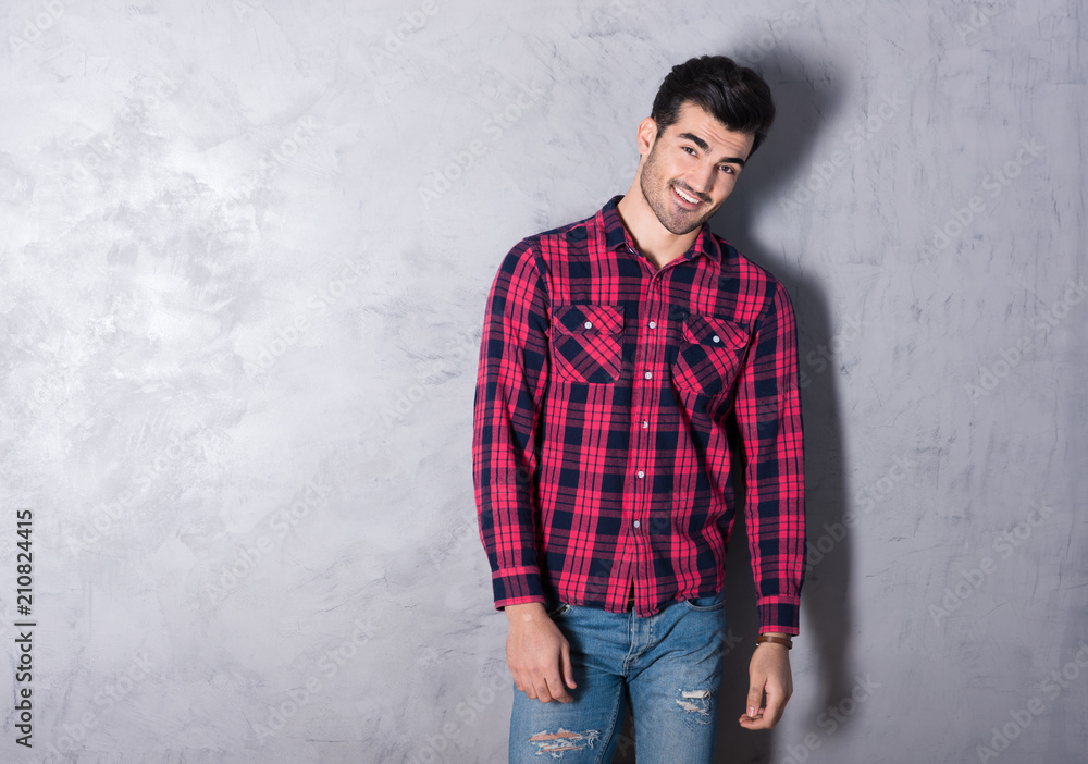 A happy handsome young man in a red checkered shirt standing in front of a grey wall in a studio.