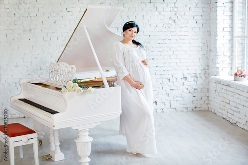 Happy pregnant girl in white dress stands near white piano photo