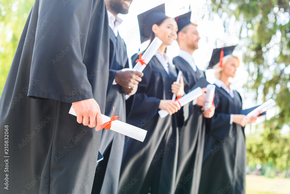 cropped shot of graduated students in capes and hats holding rolled diplomas