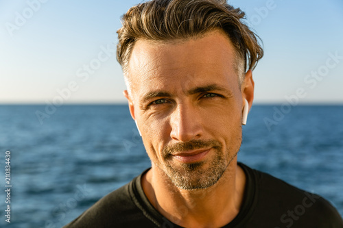 close-up portrait of smiling adult man with wireless earphones on seashore looking at camera
