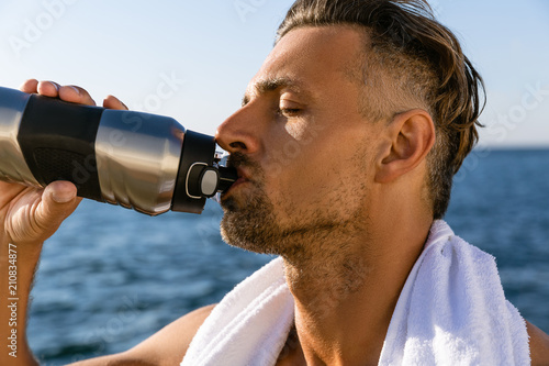 close-up shot of shirtless handsome adult man with towel on shoulders drinking water after workout on seashore
