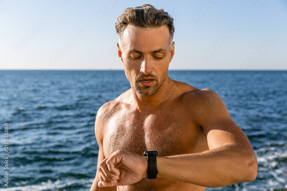 shirtless adult man looking at smart watch to check training results on seashore