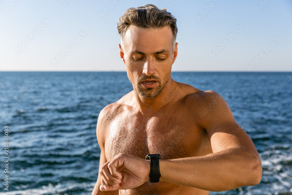 shirtless adult man looking at smart watch to check training results on seashore shirtless adult man looking at smart watch to check training results on seashore