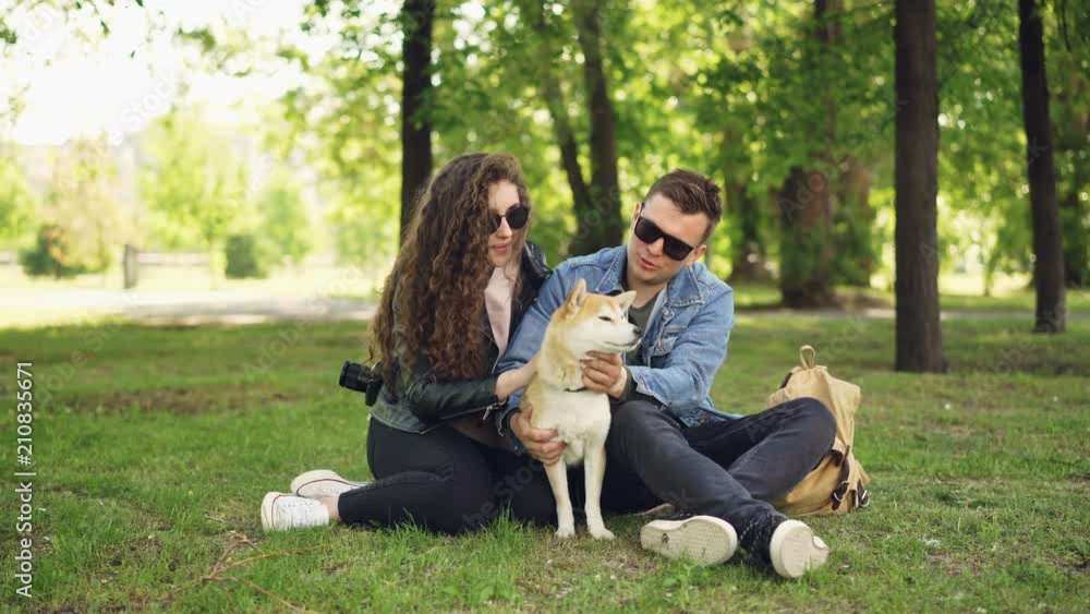 Attractive girl and her boyfriend loving dog owners are patting their pet and talking resting in the park at weekend. Happiness, nature and people concept. Attractive girl and her boyfriend loving dog owners are patting their pet and talking resting in the park at weekend. Happiness, nature and people concept.