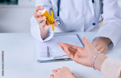 Female medicine doctor hand hold jar of pills and explain how to take pills to patient at worktable. Panacea and life save prescribing treatment legal drug store concept.